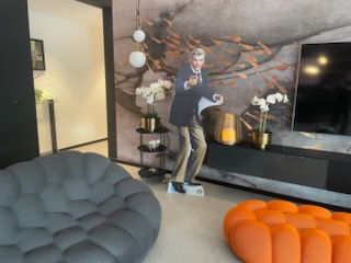Décoration intérieure avec un fauteuil gris, un pouf orange, et une silhouette cartonnée grandeur nature de James Bond pointant une arme, placée devant un mur décoratif avec fleurs blanches et télévision.