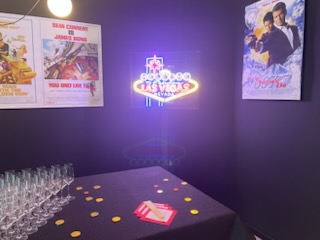 Décoration sur le thème de James Bond avec affiches de films, un néon 'Welcome to Fabulous Las Vegas', une table avec verres à champagne et jetons dorés.